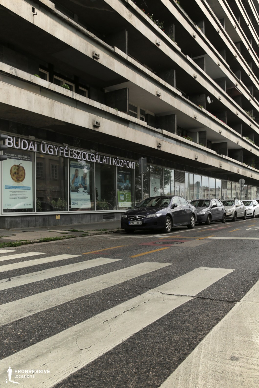This image presents a Brutalist-style building, with a stark, gray concrete facade and a series of horizontal terraces protruding from the structure. The upper levels are lined with simple concrete balconies, some decorated with potted plants, while the ground floor is dominated by large windows showcasing commercial spaces. The building's utilitarian design is emphasized by the lack of ornamentation, and its starkness is softened by the surrounding greenery and the presence of parked cars. The wide pedestrian crosswalk in front of the building adds to the urban, functional feel of the scene. This location would be ideal for film scenes set in a contemporary, industrial environment, particularly those highlighting urban landscapes, administrative spaces, or the juxtaposition of utilitarian design with everyday life. Its geometric and austere form makes it suitable for narratives involving societal or institutional settings, emphasizing functionality and modernism.