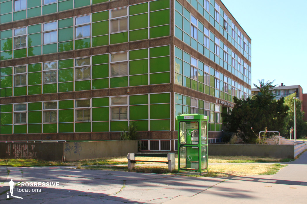 This image features the striking green glass facade of a modernist building, where large, square windows are framed by brown concrete, creating a sharp visual contrast. The building’s design is characterized by clean, geometric lines and an expansive, transparent appearance, dominated by green-tinted windows that reflect the surrounding trees. At the base, a vintage green phone booth stands next to a concrete bench, adding a touch of nostalgia to the otherwise contemporary scene. This location, with its bold geometric design and subtle urban decay, would be perfect for film scenes requiring a backdrop of institutional or public spaces, such as a hospital, government building, or office complex. The juxtaposition of modernist architecture with retro elements offers a visually rich setting for narratives focused on urban environments, government-related scenes, or the intersection of past and present in city life.