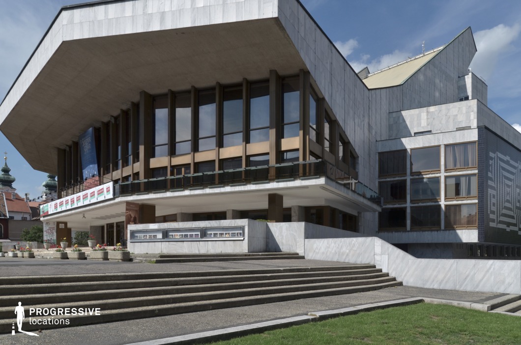 The image showcases the striking exterior of the Gyor National Theatre, a prime example of modernist architectural design. The building features sharp, angular lines, with a massive overhanging roof supported by steel beams and a combination of marble and glass on the lower levels. The prominent use of large windows creates an open, airy feel, while the imposing steps leading to the entrance enhance its monumental presence. The building's aesthetic is both contemporary and timeless, with a clean and bold design, making it ideal for scenes set in artistic or cultural environments. Its architectural grandeur, combined with the surrounding open space, provides a cinematic setting perfect for performances, public events, or urban scenes in films depicting cultural hubs or historical moments. The contrast between the theatre's modern elements and the traditional architecture in the background adds depth and character to the scene.