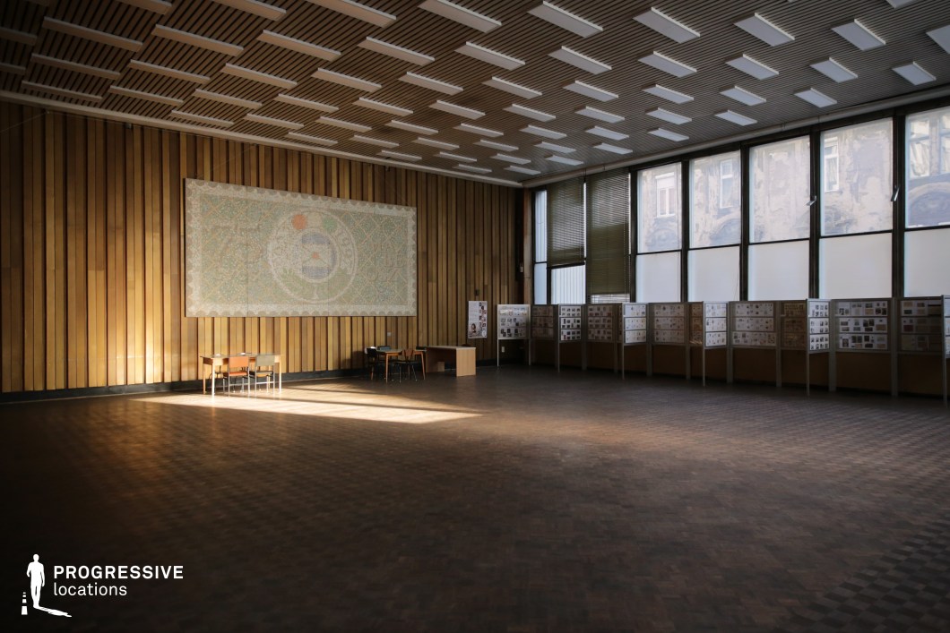 The image captures a spacious and minimalistic hall, illuminated by soft natural light filtering through large windows. The walls are adorned with wooden paneling, and the room features a tiled floor that enhances its warmth and openness. A central mosaic artwork adds an artistic focal point to the space, contrasting with the simplicity of the surroundings. On the left side, exhibition panels display various materials, creating a versatile setting for different purposes. The room's quiet and subdued aesthetic, combined with its ample natural light, makes it an ideal location for scenes that require a contemplative or artistic environment, such as quiet meetings, art exhibitions, or educational settings. Its spaciousness lends itself well to wider shots or sequences that explore the atmosphere of architectural design and space.