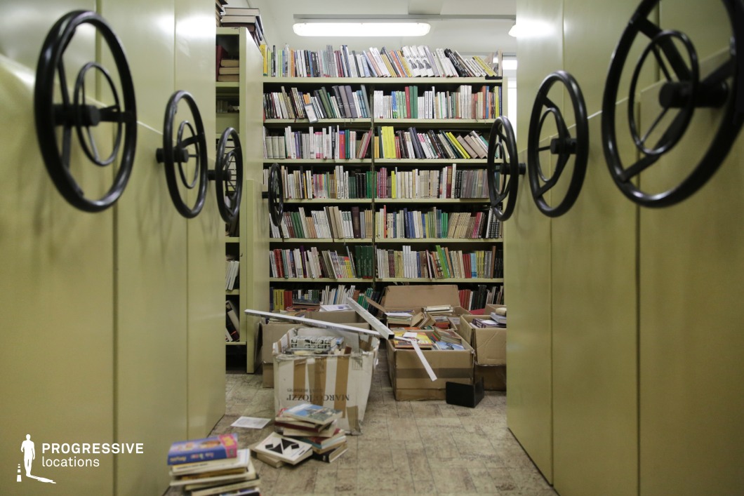 This image features a library space filled with large green shelves, each equipped with mechanical wheels for moving the units. The shelves are packed with an array of books, mostly hardcovers, arranged in a colorful and organized fashion. However, the floor is scattered with piles of disordered books, cardboard boxes, and loose papers, indicating either a recent move or an ongoing organizational process. The lighting highlights the contrasts between the neat books and the cluttered floor, offering a sense of abandonment and a busy, somewhat chaotic environment. This location would be ideal for filming scenes in settings such as a forgotten or hidden archive, a secret meeting place, or an investigative scene involving research. The mechanical bookshelves add an industrial, utilitarian atmosphere, making the space suitable for scenes requiring an aesthetic of old government-run institutions or a retro Soviet-era setting.