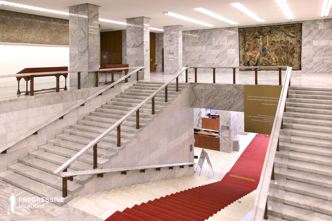 The image showcases an elegant and spacious marble-clad atrium featuring a large, central staircase that splits into two symmetrical flights. The pristine white marble floors and stair treads complement the polished wooden railings, creating a sense of luxury and grandeur. The staircase is adorned with a rich red carpet runner that contrasts beautifully against the marble. At the top, a large woven tapestry adds a touch of historical depth to the modern, minimalist architecture. The open, airy feel of the space is enhanced by the clean lines, warm lighting, and high ceilings, with further design elements such as the large marble columns supporting the upper floors. This type of location would serve well for scenes in period dramas, political thrillers, or scenes of high-society gatherings. The grand staircase and the overall design exude power and opulence, making it a perfect fit for scenes involving important decisions, meetings, or even symbolic moments in a narrative.