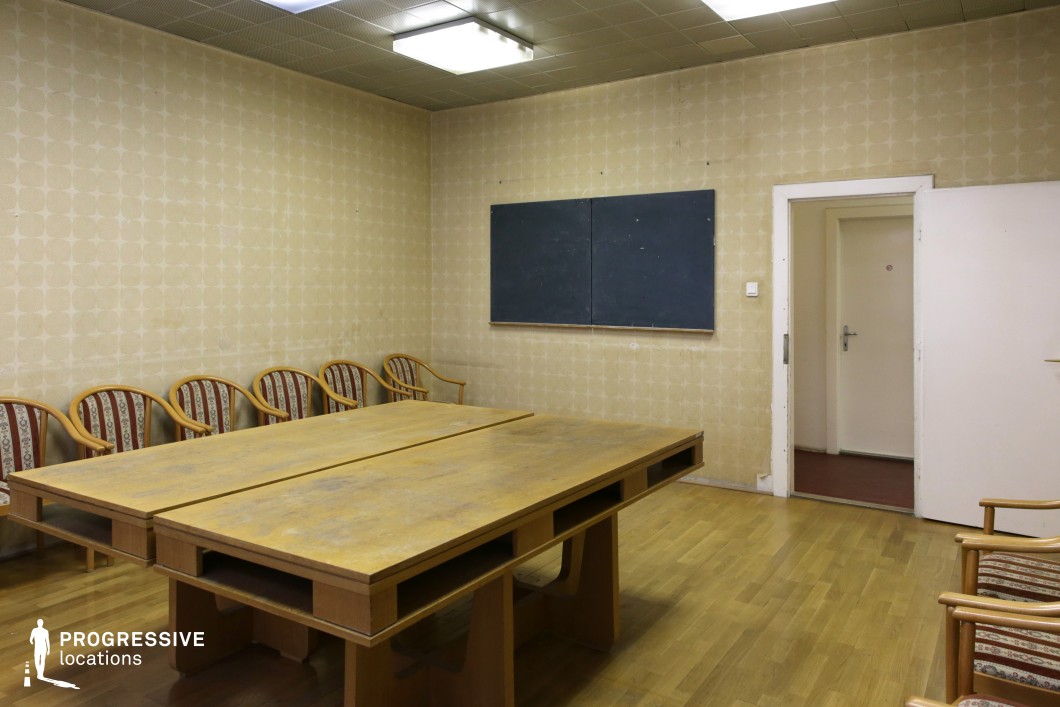 This vintage-style meeting room features a nostalgic mid-20th-century aesthetic with muted beige wallpaper adorned with geometric patterns, evoking a classic office ambiance. The room is dominated by a large, worn wooden conference table with built-in compartments, hinting at past bureaucratic functions. Surrounding it, a series of elegant wooden-framed chairs with striped and floral-upholstered backs contribute to the retro atmosphere. The flooring consists of warm-toned parquet, complementing the subdued color palette, while the ceiling is lined with square acoustic tiles, integrating simple yet functional overhead fluorescent lighting. A black chalkboard mounted on the wall, now blank and slightly worn, suggests a previous use for meetings or instructional purposes. A white-framed open doorway leads into an adjacent space with a contrasting red-tiled floor, adding depth to the composition. The room’s slightly aged appearance, with subtle marks of wear on surfaces, enhances its authenticity as a preserved historical office environment. This setting is ideal for period film productions, evoking bureaucratic or institutional themes from the past.