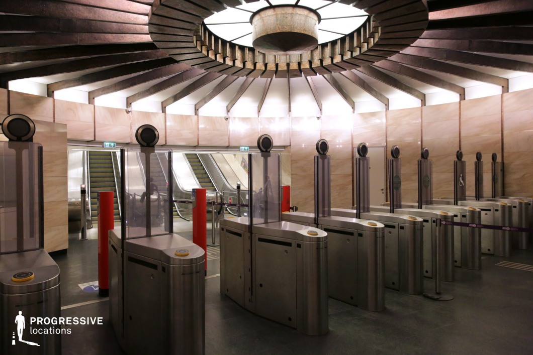 This striking metro entrance features a futuristic, almost cinematic aesthetic with a symmetrical arrangement of modern ticket barriers leading to escalators. The dominant material palette includes brushed stainless steel, polished stone, and glass, enhancing the sleek, high-tech atmosphere. The ceiling is a key architectural element, with radial, ribbed metal beams converging at a central illuminated fixture, creating a dramatic, almost sci-fi visual effect. The surrounding walls are clad in warm beige-toned panels that contrast subtly with the cool metallic elements. The gated entrance, equipped with contactless payment systems, adds to the ultra-modern functionality of the space. Red bollards break the monochromatic scheme while also guiding movement. The depth of the image, accentuated by the receding escalators and the controlled lighting, makes this an ideal location for film productions requiring a high-tech, dystopian, or urban transit setting.