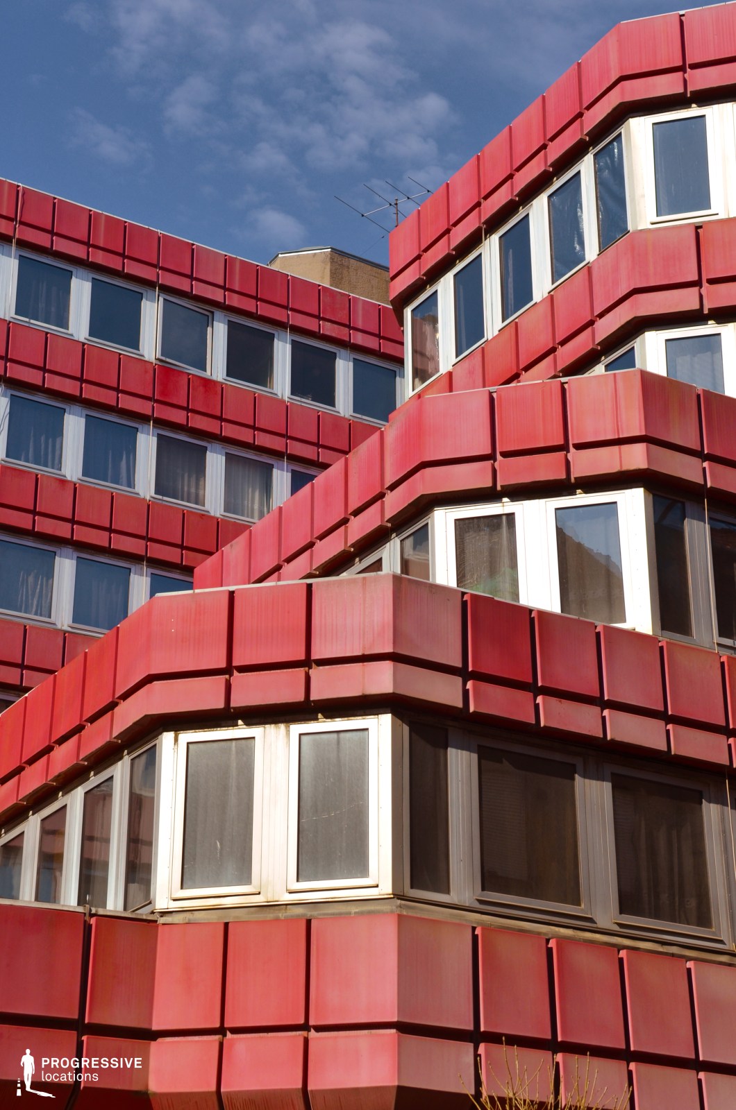 This striking building showcases a bold geometric façade, featuring modular, stacked sections clad in deep red metal panels. The angular design creates a dynamic, layered effect, with staggered balconies and protruding window sections that enhance its futuristic and Brutalist-inspired aesthetic. The building’s exterior is defined by repetitive rectangular elements, forming a rhythmic pattern that emphasizes structure and form. The contrast between the red paneling and the white-framed windows adds visual depth, while the slightly weathered surfaces give the structure an authentic, lived-in character. This location is well-suited for cinematic and creative projects requiring a retro-futuristic or Soviet-era architectural backdrop. Its dramatic geometry, combined with strong color contrasts, makes it ideal for visually compelling urban scenes or sci-fi-inspired settings.