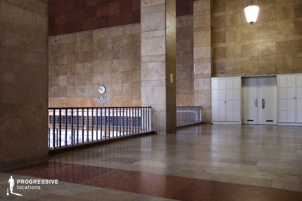 This grand transit hall exudes a mid-century monumental aesthetic, characterized by its towering marble-clad walls, high ceilings, and a timeless sense of scale. The polished stone flooring, in a mix of beige and reddish-brown hues, enhances the space’s dignified and institutional atmosphere. A key focal point is the clock mounted on the stone wall, accompanied by a set of emblematic wings, reinforcing the hall’s function as a major transportation hub. The dark metal railing frames the mezzanine level, offering a vantage point over the lower level where scheduling boards and additional transit infrastructure are visible. Large, paneled double doors in stark white contrast with the surrounding warm-toned stone, adding to the imposing yet functional character of the space. A singular pendant light fixture contributes to the sense of formality and structure. This setting is ideal for period dramas, noir-inspired cinematography, or scenes requiring an institutional, bureaucratic, or transport-related backdrop.