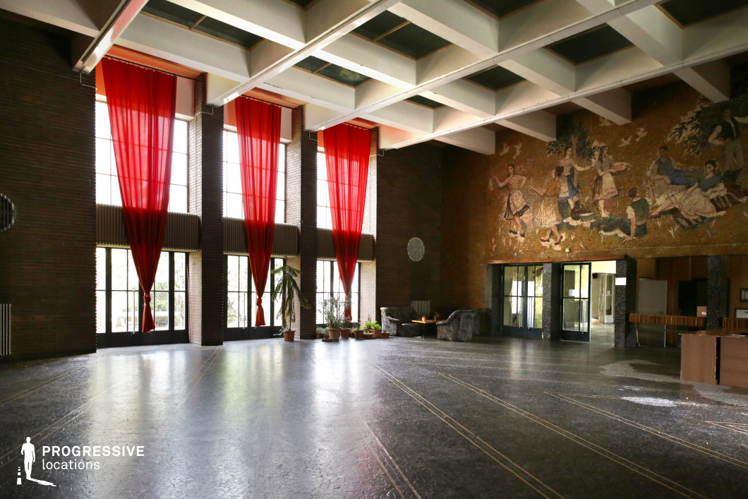 This expansive hall exudes a grand yet nostalgic mid-century aesthetic, strongly influenced by socialist-era architecture. The space is defined by towering floor-to-ceiling windows adorned with striking red sheer curtains that diffuse natural light, creating an atmospheric glow. The coffered ceiling structure adds depth and rhythm to the space, enhancing its monumental character. A key focal point is the large, intricately detailed mosaic mural on the far wall, depicting an idealized pastoral scene in the classic Socialist Realist style. The warm-toned tesserae contrast with the darker, polished floor, which features subtle linear inlays that reinforce the structured elegance of the room. Functional yet decorative elements, such as vintage lounge seating and potted plants, soften the formality of the hall. This location is ideal for period film productions, evoking themes of political grandeur, cultural institutions, or historical nostalgia, while its dramatic lighting and strong architectural features lend themselves to striking cinematography.