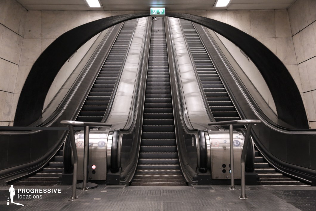 This metro station features a dramatic and futuristic escalator design, characterized by its symmetrical composition and bold, curved arch framing the ascent. The three parallel escalators, made of sleek metallic surfaces, extend upwards, creating a sense of depth and movement. The surrounding walls are clad in light-colored stone, contrasting with the dark, industrial aesthetic of the escalator structure. The black arching element enhances the cinematic appeal of the space, resembling a portal or gateway, making it an ideal setting for sci-fi, dystopian, or high-tech urban scenes. The reflective surfaces and minimalistic lighting contribute to a sterile yet dynamic atmosphere. This location is perfect for productions requiring a modern underground transit environment with a striking visual impact.