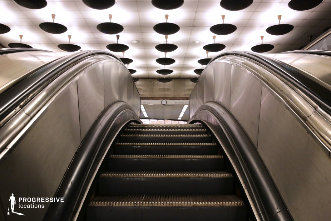 This metro station interior blends retro and futuristic aesthetics, featuring a gently curving escalator with polished metal side panels that reflect ambient light. The perspective emphasizes the smooth, flowing lines of the structure, leading toward the station’s central clock in the background. The most striking design element is the ceiling, adorned with circular black panels suspended from white tiles, creating a dramatic geometric pattern. These disc-like structures contribute to a visually dynamic and almost sci-fi atmosphere, enhancing the station’s architectural uniqueness. The subdued color palette, metallic surfaces, and symmetrical layout make this an ideal filming location for retro-futuristic, dystopian, or urban transit-themed scenes. The space’s controlled lighting and strong visual depth further enhance its cinematic potential.