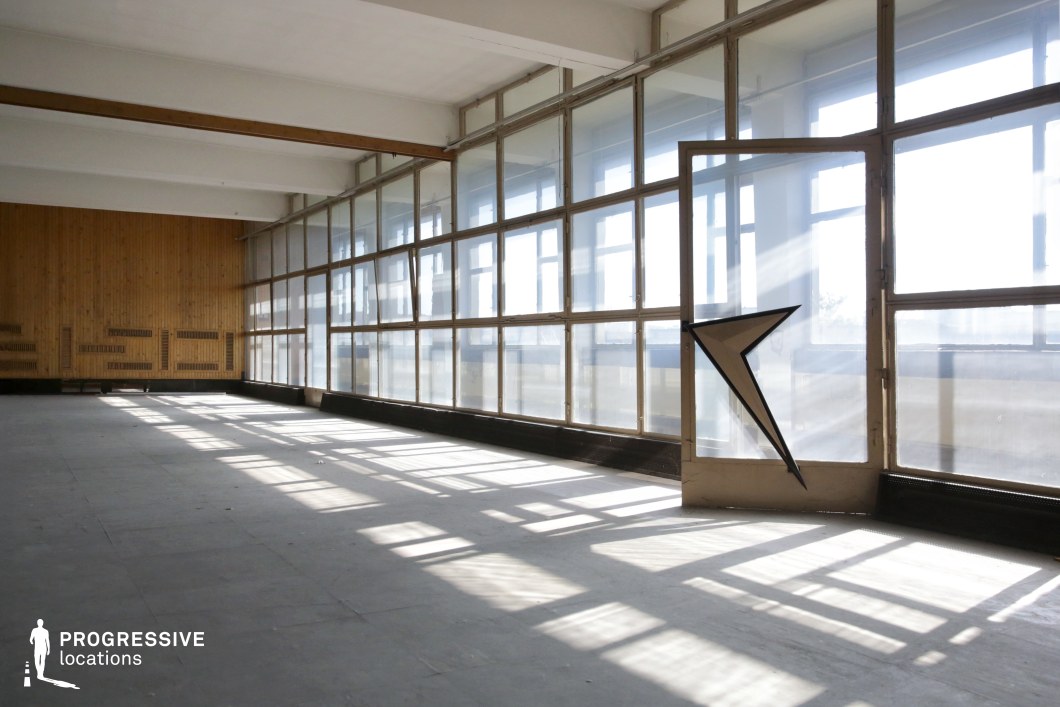 This expansive, open-plan space features a striking glass wall that allows natural light to flood the interior, casting dramatic geometric shadows across the floor. The large grid of translucent and transparent window panels gives the room an industrial yet minimalist character, enhancing its raw and unpolished aesthetic. One of the most distinctive elements is the partially shattered glass panel with a sharp, angular break, adding an element of visual tension. The far wall is clad in vertical wooden paneling, bringing warmth and texture to the otherwise stark environment. Exposed ceiling beams further emphasize the functional, utilitarian design of the space. This location is ideal for creative productions requiring an industrial, modernist, or abandoned aesthetic. The interplay of natural light and strong architectural lines makes it particularly well-suited for fashion editorials, conceptual photography, or cinematic scenes that highlight contrast between structure and decay.