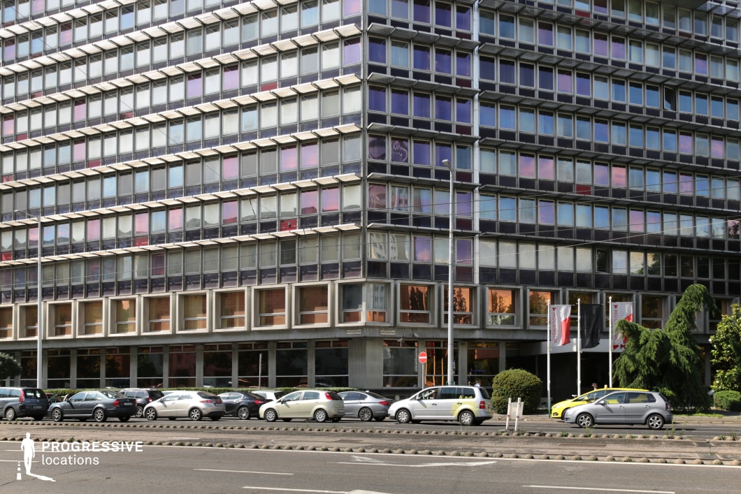 This striking mid-century modern office building features a distinctive grid-like façade composed of modular glass panels framed by dark metal elements. The reflective surfaces create a dynamic interplay of colors, with tinted and mirrored sections producing a gradient of purples, blues, and warm golden tones, depending on the angle of light. The lower section of the structure is characterized by an open, recessed ground floor with large, floor-to-ceiling windows that add transparency and depth to the design. The rigid, repetitive geometry of the building showcases a classic international style, emphasizing functionality and uniformity. The urban setting, complete with parked cars and flagpoles, reinforces the location’s identity as a corporate or institutional hub. This architectural backdrop is ideal for film productions requiring a period-accurate government or corporate setting, evoking a late 20th-century bureaucratic or financial atmosphere.