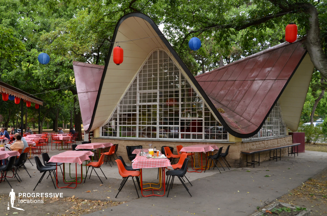This charming mid-century modern café features a distinctive undulating roofline, giving it a sculptural and dynamic appearance. The expressive architecture, with its sweeping curves and large grid-like glass façade, creates an open and inviting atmosphere, seamlessly blending the indoor space with the surrounding greenery. The outdoor seating area, set under mature trees, consists of colorful plastic chairs and red-checkered tablecloths, adding a nostalgic retro feel to the setting. The use of raw stone at the building’s base grounds the structure, while the aged metal roofing enhances its vintage character. The hanging lanterns further contribute to the playful, welcoming ambiance. This unique location is ideal for film productions or photoshoots requiring a nostalgic yet timeless European outdoor café aesthetic. Its eye-catching roof design and relaxed setting make it perfect for capturing mid-century leisure culture or contemporary adaptations of retro-futuristic design.