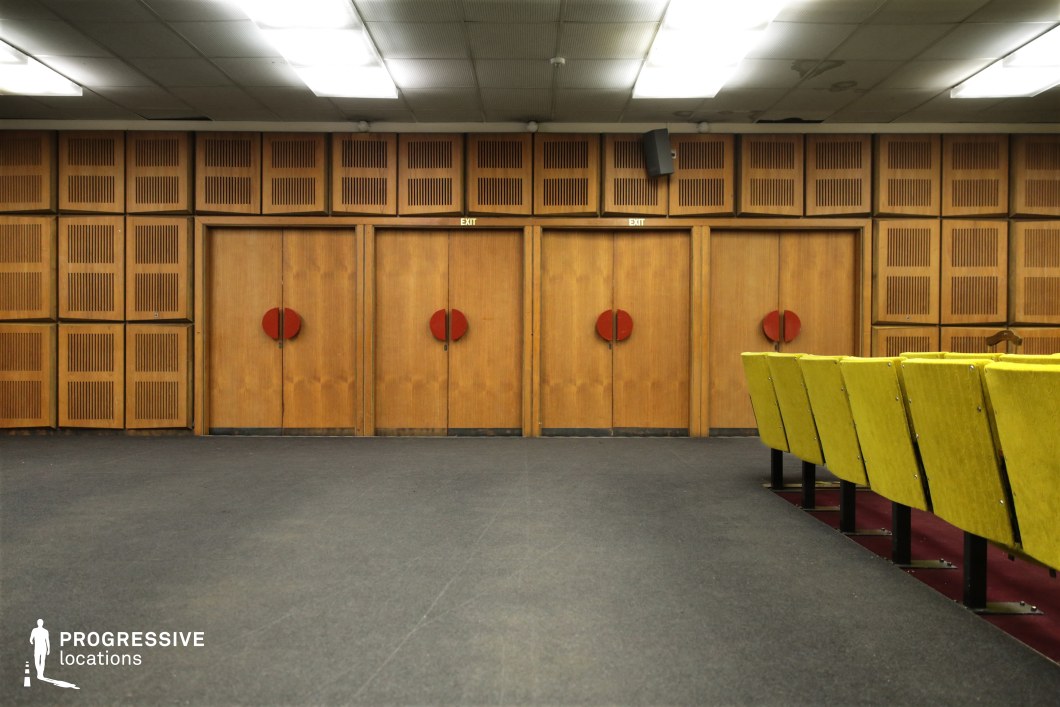 This mid-century conference hall features a warm, retro aesthetic, dominated by wooden paneling with geometric ventilation cutouts, creating a textured and uniform backdrop. The symmetrical design of the space is reinforced by three large wooden doors, each adorned with distinctive red circular handles, adding a bold visual contrast. The seating arrangement consists of rows of mustard-yellow upholstered chairs with a slightly worn texture, set against a deep red carpeted floor section, reinforcing the vintage atmosphere. The drop-ceiling, fitted with fluorescent lighting, completes the utilitarian yet character-rich ambiance. This location is well-suited for film productions requiring a period-accurate institutional or bureaucratic setting, evoking themes of mid-century government offices, academic lecture halls, or state-run cultural institutions. Its muted color palette and distinctive design details make it an ideal backdrop for both retro and minimalist visual storytelling.