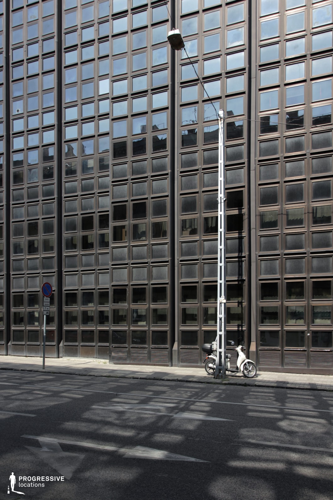 This striking modernist building features a dark, grid-like glass facade with repetitive rectangular windows, creating a structured and almost hypnotic visual effect. The reflective surfaces capture distorted glimpses of the surrounding cityscape, adding depth and movement to the rigid architectural form. In the foreground, the street is bathed in a complex play of light and shadow, as sunlight filters through an unseen structure, casting angular patterns onto the asphalt. A lone streetlamp and a parked scooter provide a sense of scale and urban solitude, emphasizing the minimalistic and somewhat dystopian aesthetic of the scene. This location is ideal for cinematic productions requiring a corporate, bureaucratic, or even futuristic setting. The contrast between the rigid, industrial architecture and the organic interplay of reflections makes it particularly well-suited for noir, cyberpunk, or architectural photography projects.