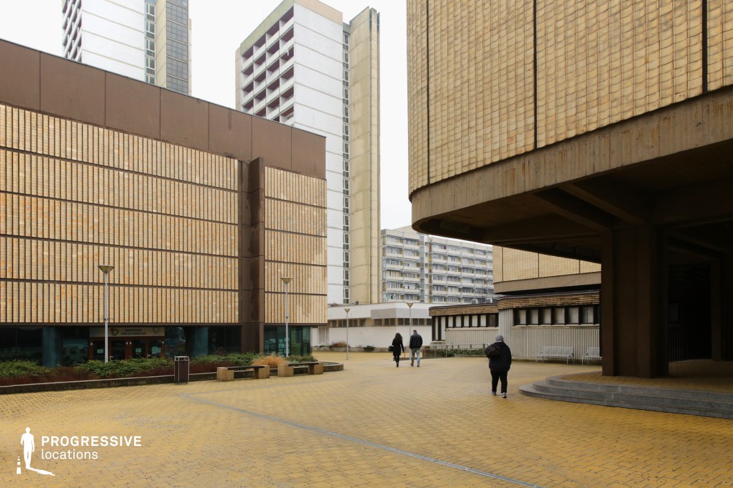 This expansive urban plaza is framed by imposing Brutalist architecture, featuring large, textured concrete and brick-clad facades in earthy yellow and brown tones. The structured, repetitive patterns on the buildings emphasize the raw, functional aesthetic of mid-to-late 20th-century institutional design. Overhanging concrete slabs supported by thick columns add to the monumental scale of the structures. The ground level is paved with yellow bricks, creating a uniform visual texture that contrasts with the verticality of the surrounding buildings. Sparse vegetation and modernist street lamps contribute to the stark, utilitarian atmosphere. In the background, high-rise residential towers with minimalist white facades reinforce the setting’s dense, planned urbanism. This location is ideal for film productions or photography projects requiring a retro-futuristic, dystopian, or Cold War-era setting. Its Brutalist forms and vast open space lend themselves to scenes depicting institutional authority, urban isolation, or architectural exploration.