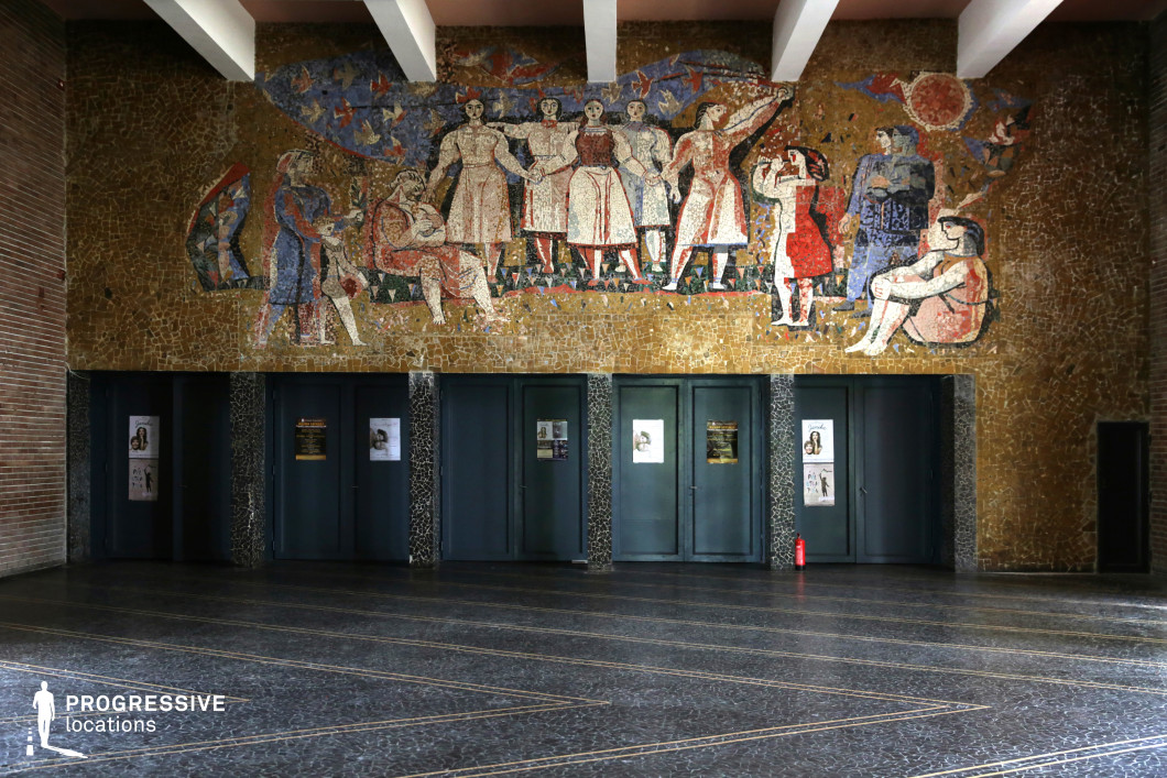 This grand interior space is dominated by a striking Socialist Realist mosaic mural, depicting an idealized vision of collective strength, labor, and social unity. The artwork, composed of warm golden hues with accents of red, blue, and white, features stylized human figures engaged in various communal activities, set against a backdrop of abstract geometric patterns and natural elements. Beneath the mural, a row of dark green doors provides an institutional aesthetic, framed by black terrazzo-clad columns. The polished stone flooring features embedded linear patterns, reinforcing the structured, authoritative atmosphere of the hall. The exposed ceiling beams add to the monumental scale of the space, enhancing its mid-20th-century architectural character. This location is ideal for film productions or photography projects requiring an authentic Soviet-era or institutional setting. The powerful visual narrative of the mosaic, combined with the rigid symmetry of the architecture, makes it a compelling backdrop for historical, political, or dystopian themes.