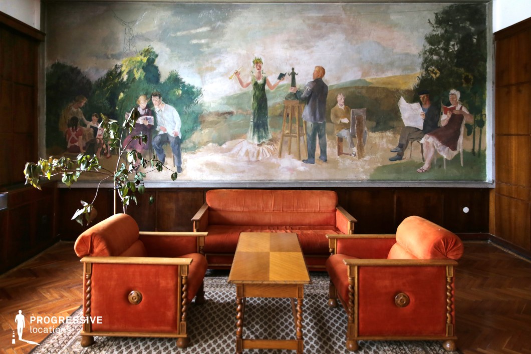 This vintage lounge space is characterized by warm wood paneling, deep red-orange upholstered seating, and a striking Socialist Realist mural that spans the wall. The painting, executed in soft, muted tones, depicts an idealized pastoral scene where people engage in artistic and intellectual activities, evoking a utopian vision of creativity and learning. The furniture, featuring carved wooden details and plush velvet upholstery, reinforces the mid-century aesthetic. A patterned carpet adds texture, while the parquet flooring enhances the nostalgic charm. The presence of a leaning potted plant introduces a subtle contrast to the structured arrangement. This location is ideal for film productions or photography projects requiring an authentic historical setting, evoking mid-20th-century intellectual salons, cultural institutions, or government reception spaces. The carefully preserved elements make it a perfect backdrop for period dramas or artistic narratives.