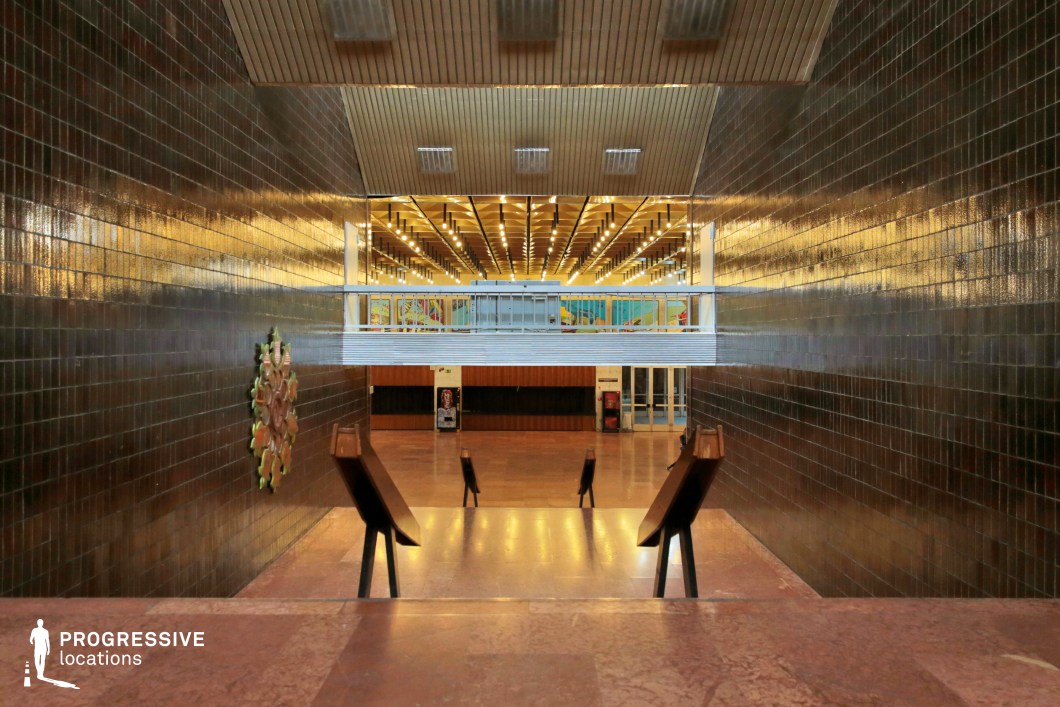 This striking university interior features a wide, descending staircase leading to an open hall bathed in golden light. The walls are clad in dark, glossy tiles that reflect the linear ceiling lights, creating an almost cinematic atmosphere. The contrast between the lower, dimly lit foreground and the illuminated upper space enhances the sense of depth and perspective. The hall’s ceiling showcases a structured grid of hanging lights, adding rhythm and symmetry to the design. Decorative elements, such as a wall-mounted ceramic artwork and mid-century modern wooden reading stands, reinforce the retro academic aesthetic. A distant mural adds a subtle pop of color, hinting at the building’s cultural or educational purpose. This location is ideal for film productions or photography projects requiring a mid-century institutional setting. The combination of rich textures, dramatic lighting, and functionalist design makes it perfect for academic, bureaucratic, or even dystopian-themed narratives.