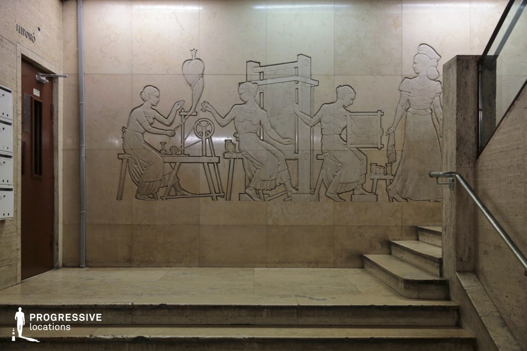 This institutional interior features a beautifully carved bas-relief mural set into a polished marble wall, depicting a Socialist Realist scene of women engaged in traditional textile production. The figures, rendered in a classical style, emphasize labor, craftsmanship, and collective effort, aligning with mid-20th-century ideological aesthetics. The surrounding space is characterized by muted beige and cream-colored stone, with a simple yet imposing staircase leading upward. A metal handrail and a functional brown door contrast with the refined elegance of the wall relief. The clean lines and smooth surfaces contribute to the formal, bureaucratic atmosphere of the setting. This location is ideal for film productions or photography projects requiring an authentic Soviet-era institutional or educational environment. The combination of grand yet restrained architecture and ideological artistic elements makes it a compelling backdrop for historical, political, or documentary-style narratives.