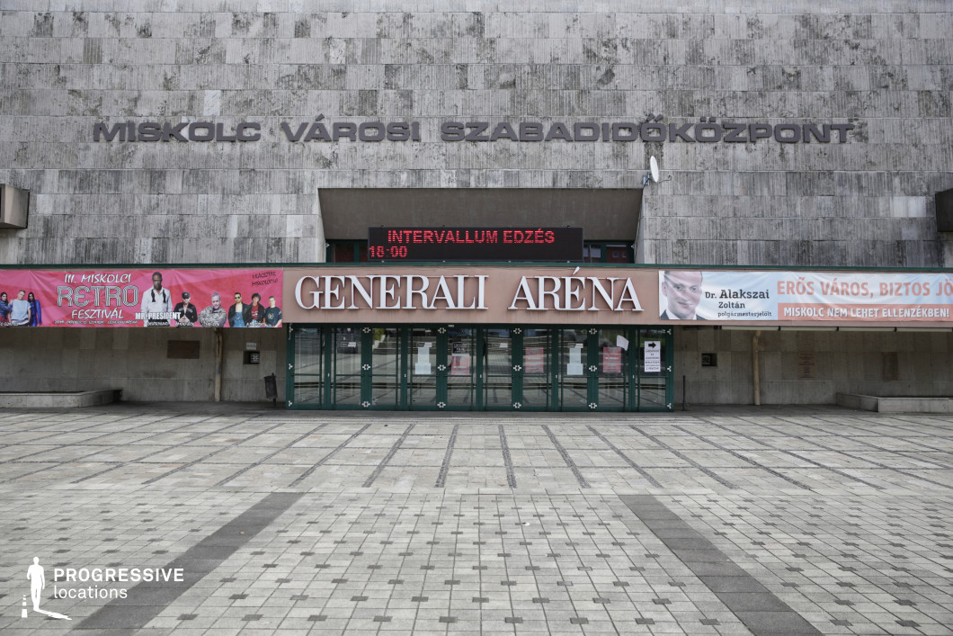 This imposing Brutalist-style sports and event arena features a massive stone-clad facade with bold, industrial lettering, emphasizing its utilitarian and monumental aesthetic. The textured grey stone exterior is punctuated by a series of modern glass entrance doors, sheltered under a horizontal awning displaying signage and advertisements. A large digital display board above the entrance provides real-time updates on events, while banners and posters add a contemporary layer of visual contrast to the otherwise stark architectural composition. The surrounding plaza is paved with geometric tiles, reinforcing the structured, urban character of the space. This location is ideal for film productions or photography projects requiring a mid-to-late 20th-century institutional backdrop. The Brutalist design, combined with the large open plaza, makes it suitable for themes involving sports, concerts, public gatherings, or governmental architecture. Its strong geometric presence also lends itself well to dystopian or retro-futuristic visual storytelling.
