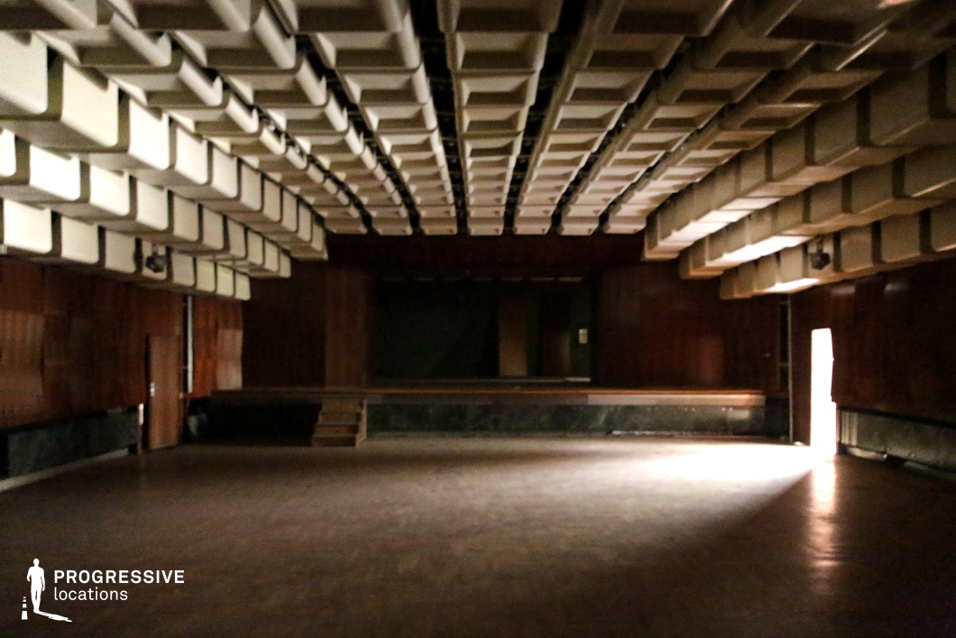 This atmospheric, dimly lit auditorium features a striking geometric ceiling design composed of protruding rectangular acoustic panels, creating a bold, sculptural effect. The space is clad in warm wood paneling, contributing to a mid-century institutional aesthetic. A small, elevated stage with a darkened backdrop suggests past performances or lectures, while the worn parquet flooring enhances the aged, nostalgic character of the venue. A single open doorway allows light to seep in, casting long shadows across the floor and heightening the cinematic quality of the space. The lack of seating and minimal furnishings adds to the abandoned or repurposed atmosphere, making it a versatile backdrop for film productions or photography projects. This location is ideal for scenes requiring a retro-futuristic, theatrical, or even dystopian setting. The combination of dramatic lighting, bold architectural elements, and a sense of isolation makes it particularly well-suited for conceptual storytelling, music videos, or experimental visual compositions.