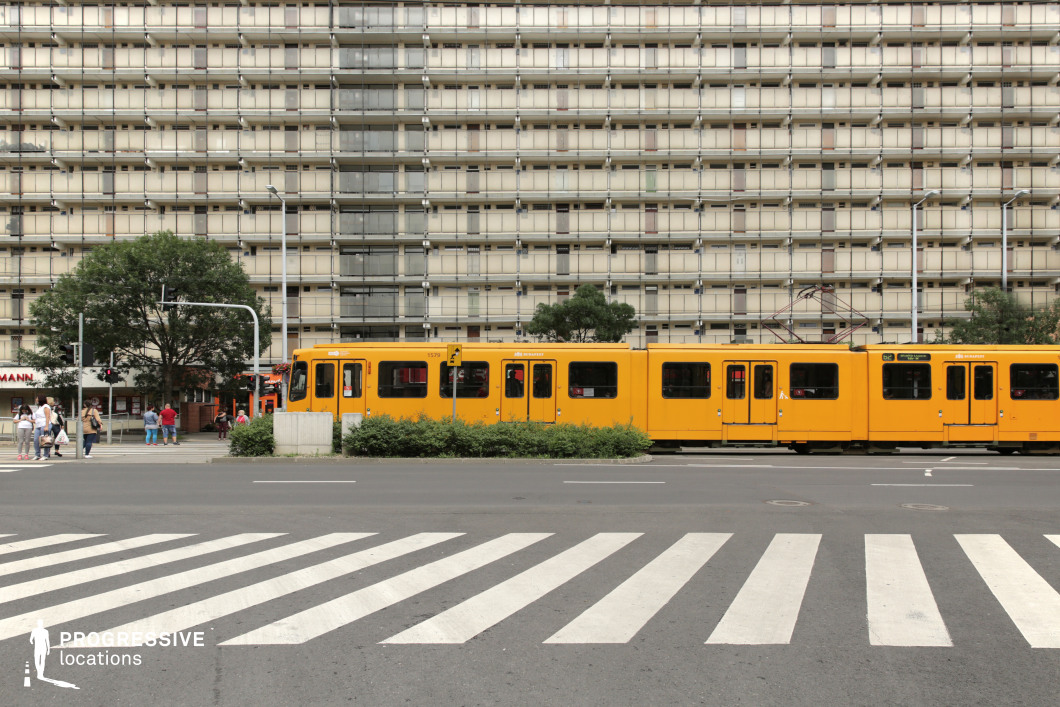 This urban streetscape captures a striking contrast between the bold yellow of a passing tram and the repetitive, grid-like facade of a massive prefabricated apartment block in the background. The structured, symmetrical design of the residential building, characterized by uniform balconies and muted beige tones, evokes a classic socialist-era architectural style. A wide pedestrian crosswalk in the foreground adds depth and movement to the composition, guiding the viewer’s eye toward the tram and the densely built environment beyond. The scene is punctuated by urban elements such as traffic lights, street signs, and pedestrians waiting at the intersection, enhancing the everyday metropolitan atmosphere. The subdued, neutral tones of the architecture emphasize the tram’s vibrant yellow, creating a dynamic yet balanced color contrast. This location is ideal for film productions or photography projects that require a visually compelling urban setting, making it perfect for themes involving public transportation, city life, or Eastern European-style modernist architecture.