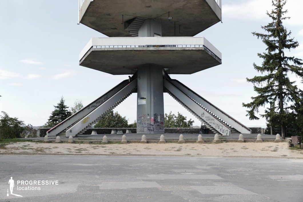 This imposing brutalist observation tower features a striking geometric structure with two massive, cantilevered platforms supported by a central concrete pillar. Two symmetrical staircases lead upwards from the ground level, creating a visually balanced composition, while an additional spiral staircase at the core allows access to the highest platform. The raw concrete surfaces, adorned with graffiti, emphasize the structure’s urban and weathered aesthetic, adding layers of texture and character. The surrounding environment consists of an open paved area with scattered greenery, punctuated by stone barriers and a few trees, reinforcing the sense of isolation and prominence of the structure. The vast sky and distant treetops provide a contrasting natural backdrop, making this location ideal for dystopian cinematography, urban exploration themes, or dramatic architectural photography.