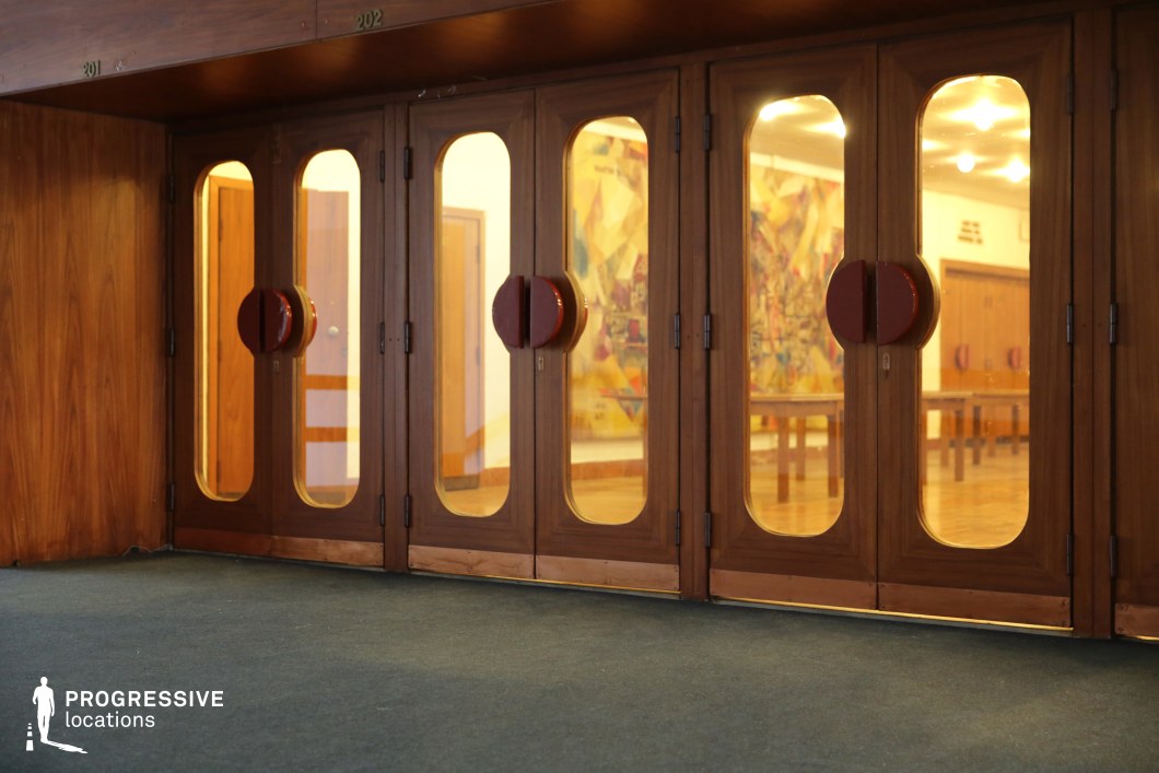 This set of elegant mid-century wooden doors features a symmetrical design with rounded glass panels framed in warm-toned wood. The distinctive circular red handles add a bold visual contrast while maintaining a cohesive vintage aesthetic. Through the glass panes, a warmly lit interior reveals a spacious hall with wooden furniture and a striking abstract mural on the far wall. The deep green carpet in the foreground enhances the retro ambiance, complementing the rich wood tones. The soft glow of ceiling lights inside creates an inviting atmosphere, making this location ideal for period-set productions, nostalgic architectural photography, or film scenes requiring a refined, historic indoor space.