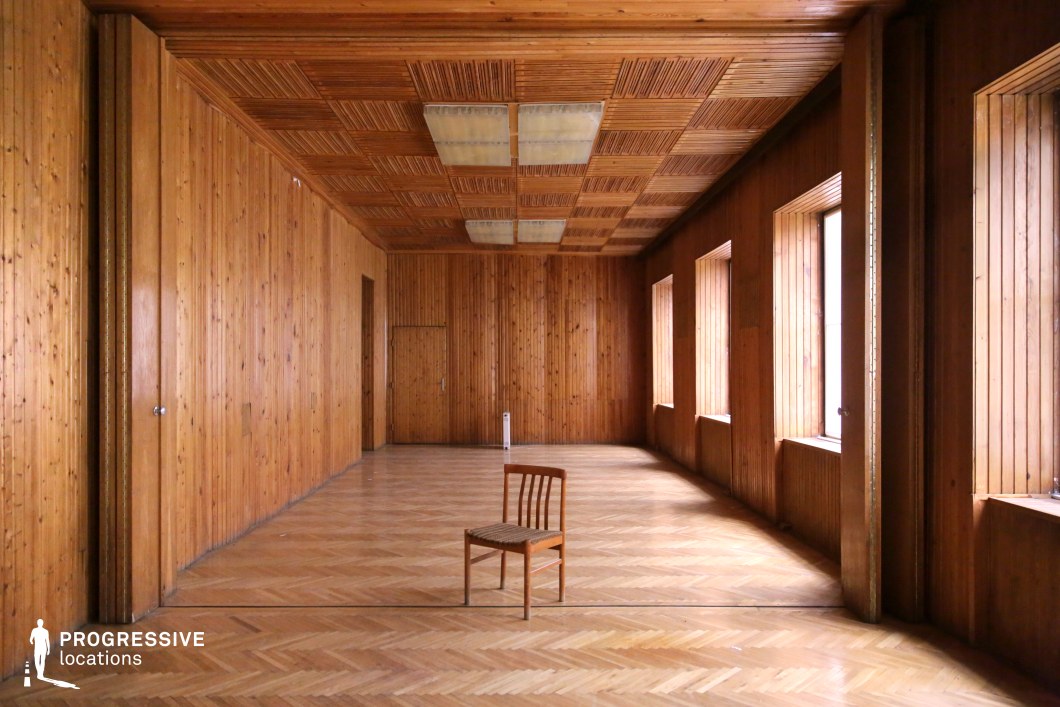 A spacious, fully wood-paneled room featuring a warm, natural aesthetic with a rhythmic repetition of geometric patterns on the walls, ceiling, and parquet flooring. The symmetrical arrangement of rectangular windows allows soft natural light to stream in, casting subtle shadows that enhance the texture of the wooden surfaces. The ceiling showcases a grid-like pattern with recessed panels, adding depth and an architectural focal point. The lone wooden chair positioned at the center of the room reinforces the minimalist and contemplative atmosphere, making this an ideal setting for artistic, conceptual, or introspective film scenes. The combination of warm wood tones and empty space offers flexibility for various creative visual compositions.