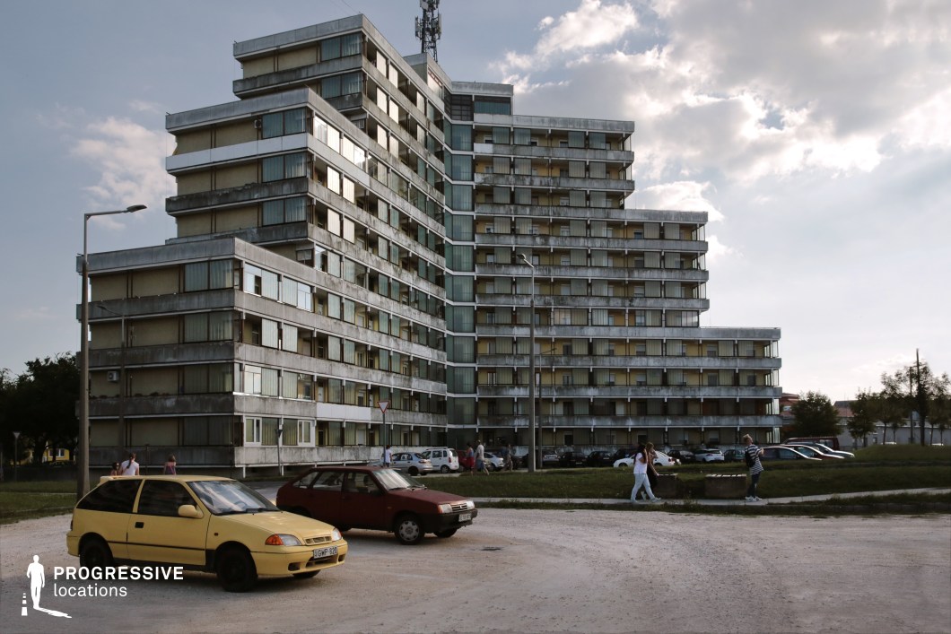 A striking example of brutalist architecture, this large-scale terraced housing complex features a stepped, geometric design with a repetitive pattern of balconies and windows. The building's exterior consists of weathered concrete and glass panels, emphasizing a raw, utilitarian aesthetic typical of mid-to-late 20th-century urban developments. The surrounding open space includes sparse greenery, a gravel parking area, and scattered vehicles, reinforcing the atmosphere of a lived-in, functional residential setting. The angular form of the building, combined with its asymmetrical facade, creates a sense of depth and scale, making it a visually compelling location for scenes depicting urban life, social realism, or dystopian narratives.