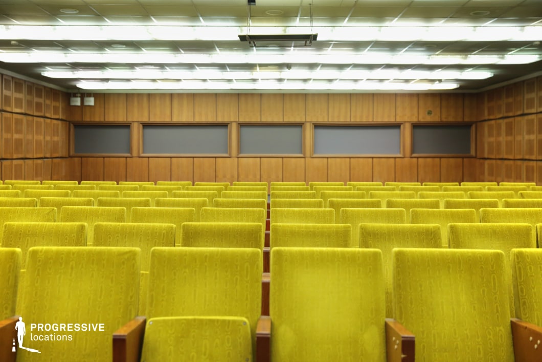 This mid-century style auditorium features rows of vibrant yellow upholstered seats with wooden armrests, arranged in a symmetrical, linear pattern that emphasizes depth and perspective. The walls are clad in warm wood paneling, creating a balanced contrast with the seating and lending a vintage character to the space. Overhead, a grid of recessed fluorescent lighting evenly illuminates the room, reinforcing the retro aesthetic reminiscent of conference halls, lecture theaters, or classic screening rooms from the late 20th century. The neutral gray panels on the back wall add a subdued, functional element, likely for acoustic or projection purposes. This location is ideal for film scenes requiring an old-school educational setting, a vintage corporate environment, or a nostalgic throwback to a past era of public institutions.