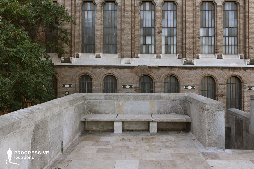 A historic urban terrace constructed from weathered limestone features an integrated stone bench, enclosed by a sturdy parapet, creating a structured yet aged aesthetic. The terrace is set against a grand brick façade with tall, arched windows, framed by stone accents, emphasizing symmetry and classical architectural influence. The combination of warm brown brickwork and pale limestone provides a subtle contrast, while the presence of a lush green tree softens the rigid geometry. The terrace’s stone flooring, laid in irregular patterns, reinforces its aged character. This space, with its enclosed yet open atmosphere, is ideal for contemplative or refined urban scenes, offering a visually compelling setting with strong architectural depth and material contrasts.