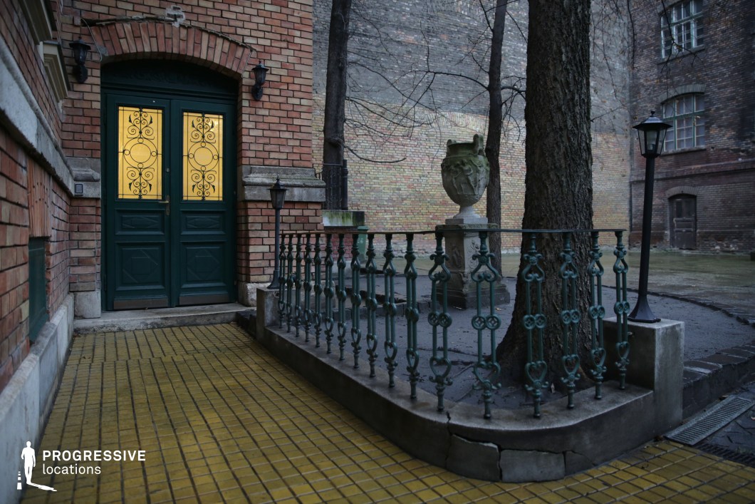 A historic brick courtyard featuring an ornate green double door with decorative wrought-iron inlays, softly illuminated from within. The entrance is framed by aged brickwork with arched detailing, adding to its classical charm. A yellow-tiled pathway leads to the door, contrasting with the dark, weathered metal railing enclosing a small raised section around a tree. The courtyard is accentuated by vintage-style street lamps and an aged stone urn on a pedestal, reinforcing the location’s historic ambiance. The surrounding walls, composed of varied brick textures, enclose the space, creating a secluded yet cinematic setting. This visually rich and atmospheric environment is ideal for period dramas, mystery scenes, or character-driven narratives.