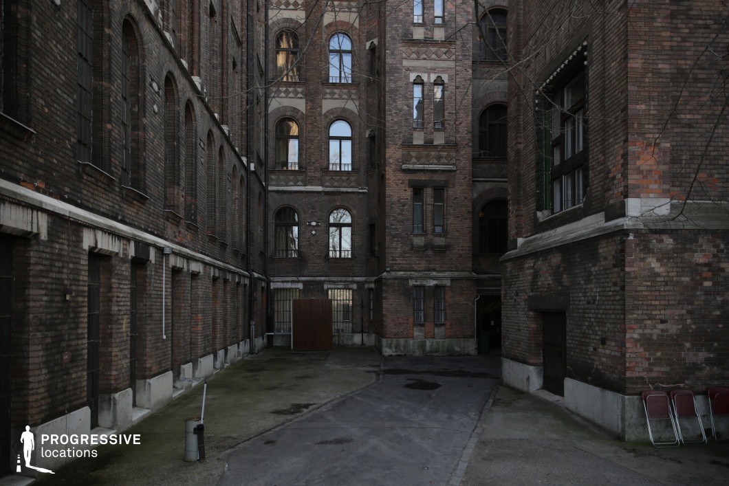 A secluded, enclosed courtyard surrounded by tall, historic brick buildings with arched windows and intricate masonry details. The aged brickwork exhibits a mix of dark and warm tones, contributing to a textured, timeworn aesthetic. The narrow, irregularly paved ground, lined with barred windows and simple structural elements, enhances the enclosed and somewhat industrial atmosphere. The repetition of arched openings and decorative brickwork adds architectural depth, while scattered utilitarian objects hint at the space’s functional past. The lack of direct modern modifications preserves its historic character, making it an ideal backdrop for period dramas, noir-inspired scenes, or urban storytelling with an atmospheric, cinematic quality.