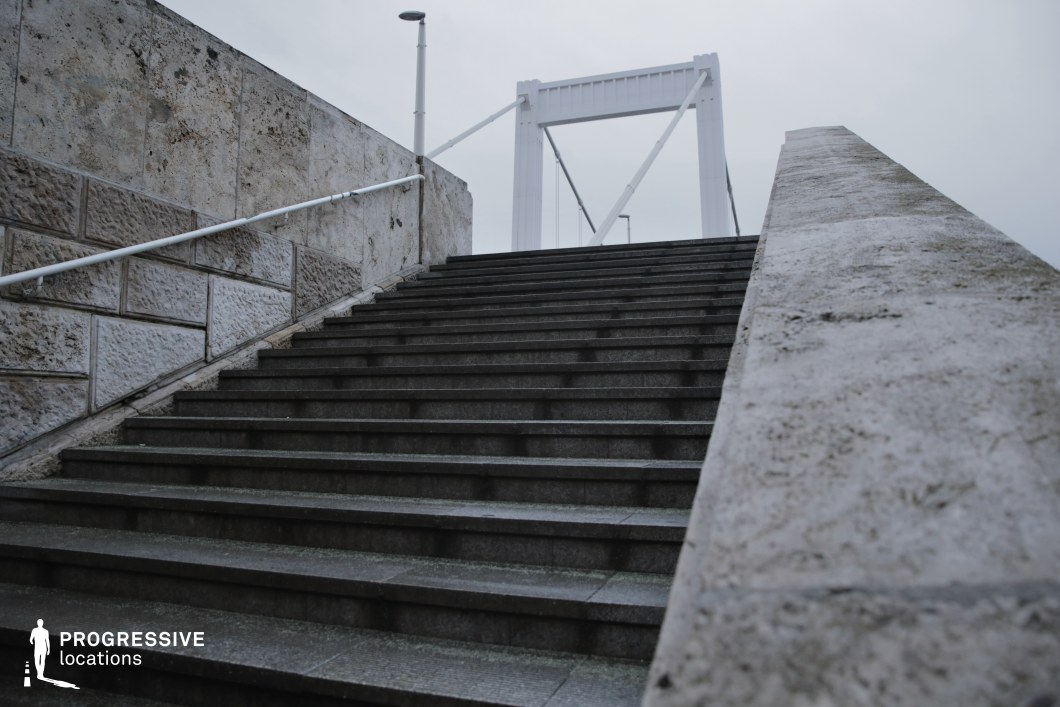 A grand stone staircase with dark, weathered steps leads towards a modern suspension bridge, framed by textured limestone walls. The structured composition emphasizes depth and perspective, guiding the eye upwards towards the geometric white steel frame of the bridge. The clean, minimalist aesthetic of the bridge contrasts with the raw texture of the stone, creating a visual dialogue between old and new architectural elements. Metal railings and lampposts add functional urban details, enhancing the structured, urban environment. This location is ideal for cinematic sequences requiring dynamic movement, symbolic ascension, or modern urban storytelling.