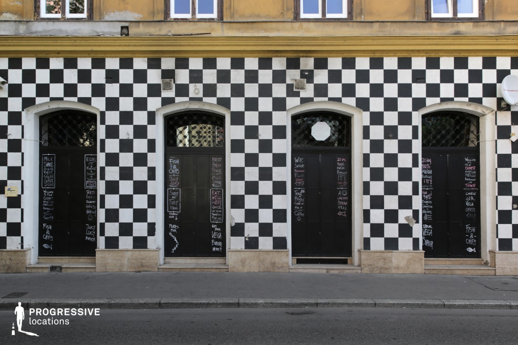 A striking black-and-white chequered building facade creates a bold, eye-catching visual identity. The symmetrical design features arched doorways with dark metal doors, adorned with handwritten chalk menus, suggesting a restaurant or café setting. Above, a contrasting yellow upper section with aged wooden window frames adds a layer of historical character. The geometric patterning gives the exterior a modern yet playful energy, while the weathered details hint at a long-established presence. This unique urban backdrop, with its mix of vintage and contemporary elements, is ideal for creative, dynamic, and alternative visual storytelling.