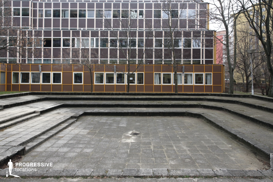 A stepped concrete courtyard unfolds in a geometric arrangement, leading to a modular, mid-century modern building with a striking grid-like facade. The structure features a combination of brown and yellow paneling with large, evenly spaced windows, creating a uniform and repetitive architectural pattern. The courtyard’s tiered layout, framed by low retaining walls, suggests a communal or educational space, possibly an outdoor amphitheater or gathering area. Leafless trees punctuate the scene, adding vertical contrast to the structured composition. The raw concrete and retro-futuristic building design make this location ideal for institutional, academic, or urban social settings in visual storytelling.