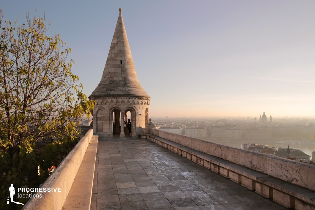 A grand limestone terrace leads to an ornate turret with arched openings, offering an expansive view of the cityscape below. The structure, with its conical roof and intricate stone detailing, evokes medieval and neo-Romanesque architectural influences. The elevated walkway, bordered by a solid stone balustrade, adds depth and perspective, guiding the eye toward the open archways that frame the skyline. The surrounding elements, including the textured stone flooring and adjacent greenery, enhance the contrast between nature and built heritage. This location serves as a stunning backdrop for period settings, historical narratives, or dramatic urban cinematography.