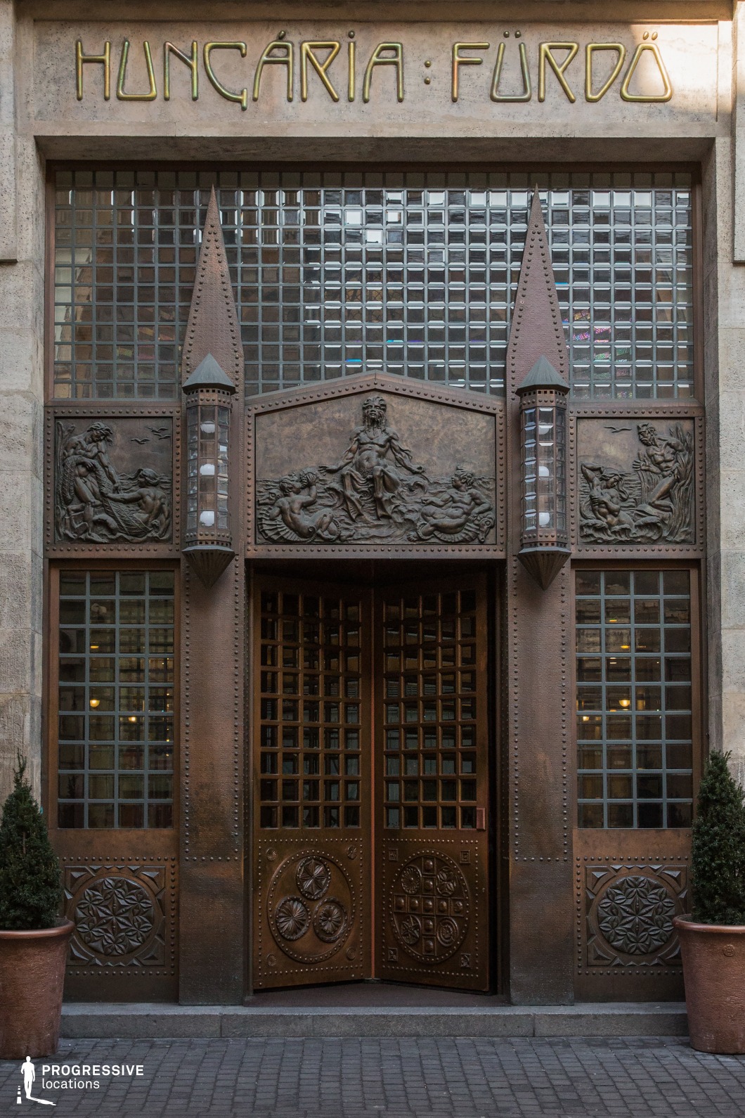 An opulent Art Nouveau entrance featuring a grand copper doorway adorned with intricate bas-relief sculptures depicting mythological water scenes. The symmetrical design is framed by two pointed decorative elements, each housing vertical glass lanterns, enhancing the structure’s architectural richness. Above, a glass-block window allows soft, diffused light to enter, complementing the warm metallic tones of the façade. The elegant, stylized lettering adds to the historical character of the location. This striking entrance, with its fusion of artistic craftsmanship and functional design, is an ideal setting for period dramas, luxury-themed visuals, or architectural heritage documentation.