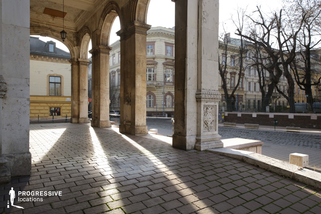A stately colonnade with intricately carved stone pillars and elegant arches frames a picturesque cityscape of historic buildings. The textured stone floor, arranged in a grid-like pattern, enhances the structured, classical ambiance. Sunlight filters through the arches, casting dramatic linear shadows that emphasize depth and perspective. The weathered architectural details suggest a rich historical context, while the surrounding urban environment, featuring ornate facades and cobblestone streets, adds to the old-world charm. This location is ideal for period dramas, architectural photography, or cinematic compositions that highlight grandeur and historic depth.