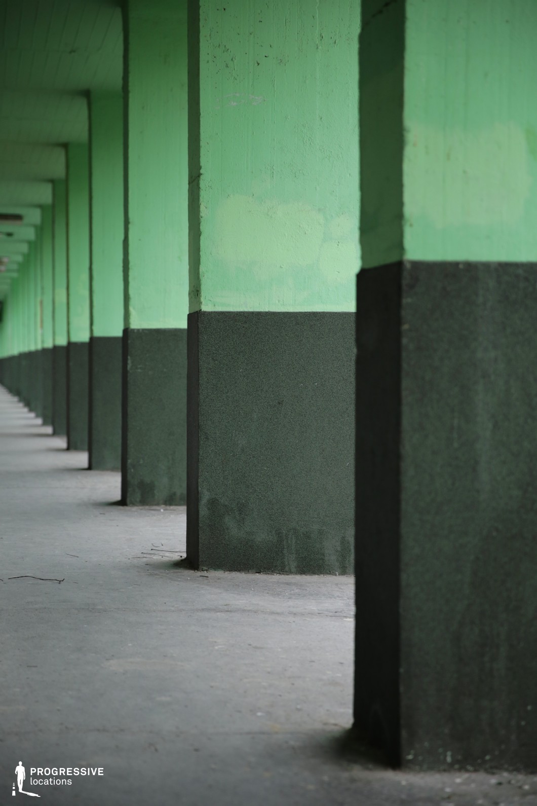 This image showcases a series of evenly spaced, massive concrete columns forming a repetitive architectural pattern. The columns are painted in a muted green hue on the upper section, while the lower half features a darker, almost black finish, creating a stark contrast. The structure extends into the distance, creating a strong sense of depth and linear perspective. The textured surfaces and subtle wear on the paint add an industrial, utilitarian character to the setting. The covered walkway suggests an urban or suburban location, possibly part of a public transportation hub, stadium, or large industrial facility. This setting is ideal for scenes requiring a raw, structured, and somewhat isolated atmosphere, offering striking symmetry and visual rhythm for cinematographic compositions.