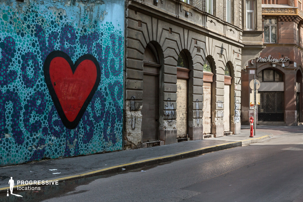 This image captures a textured urban streetscape where modern street art contrasts with historic architecture. A large graffiti mural dominates the left side, featuring a bold red heart outlined in black, set against a vibrant blue and turquoise dotted background. The adjacent building showcases a classical façade with arched doorways, decorative masonry, and aged surfaces marked by peeling paint and graffiti tags. The rolling metal shutters suggest a once-active commercial space now abandoned or repurposed. The street is narrow, lined with a sidewalk and a red fire hydrant, while the warm-toned architecture in the background adds depth and authenticity to the scene. The mix of artistic expression and weathered urban decay creates an ideal backdrop for gritty, cinematic city sequences or editorial fashion photography.