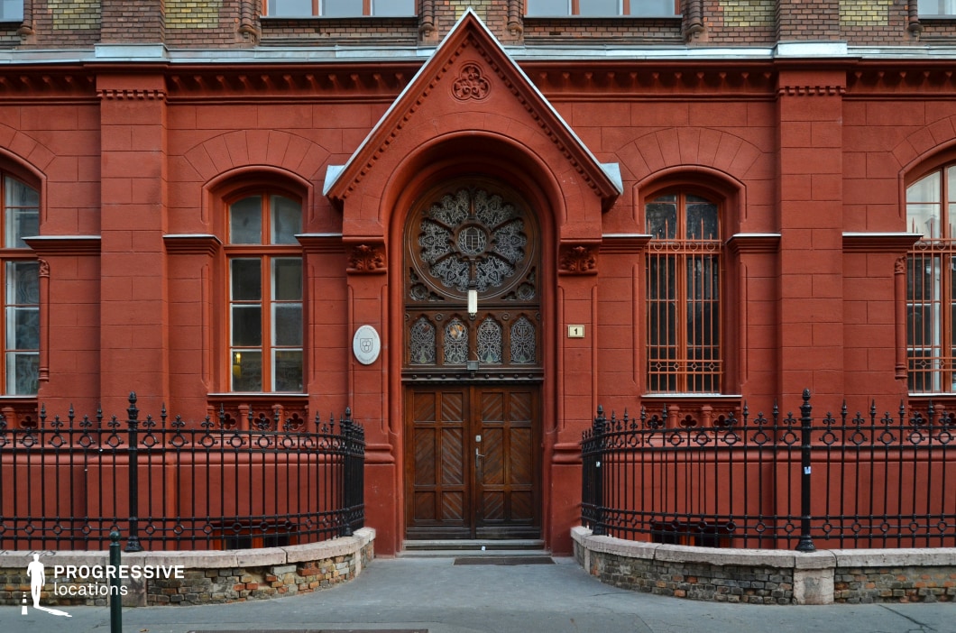 This image features a striking historical building with a deep red brick façade and intricate architectural details. The grand arched entrance is adorned with an elaborately carved wooden door, featuring wrought iron and glass inlays that add a sense of craftsmanship and elegance. Above the entrance, a decorative gable enhances the structure’s classical character. The symmetrical windows, framed by subtle brickwork patterns and secured with ornamental iron bars, contribute to the refined aesthetic. A black wrought-iron fence with intricate designs encircles the entrance, resting on a low brick and stone foundation. The mix of red brick, dark wood, and metalwork gives the scene a European, institutional feel, making it an ideal setting for historical dramas, academic-themed shoots, or refined urban storytelling.