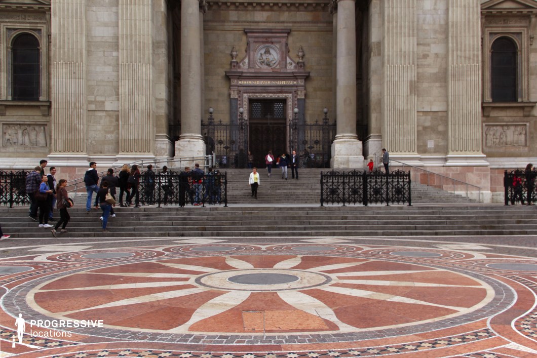 This image showcases the grand entrance of a neoclassical basilica, featuring towering fluted columns, a richly decorated tympanum, and imposing dark metal gates. The symmetrical stone façade is adorned with intricate carvings and sculptural details, reinforcing its historical and architectural significance. Leading up to the entrance is a wide, multi-tiered staircase, framed by decorative wrought-iron railings. In the foreground, a large circular mosaic with an elaborate radial pattern, composed of warm-toned stones, enhances the visual depth of the scene. The combination of classical architecture and ornamental pavement creates a striking, formal setting, ideal for historical and cultural productions or sophisticated urban visuals.
