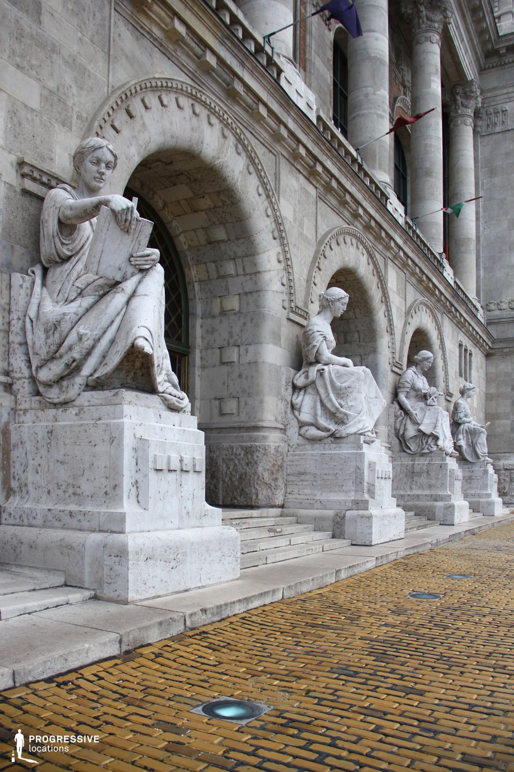 This image showcases an elegant neoclassical building with a row of intricately carved limestone statues seated on pedestals along its arched façade. The figures, draped in flowing garments, hold symbolic objects, suggesting themes of knowledge, wisdom, or the arts. The building features a richly detailed exterior with symmetrical arches, decorative moldings, and towering columns, reinforcing its institutional and historical significance. The foreground presents a paved walkway of yellow-toned cobblestones, embedded with circular in-ground lights that add a modern touch to the otherwise classical setting. The combination of grand architecture, sculptural artistry, and refined materials makes this an ideal backdrop for academic, cultural, or historical-themed visual projects.