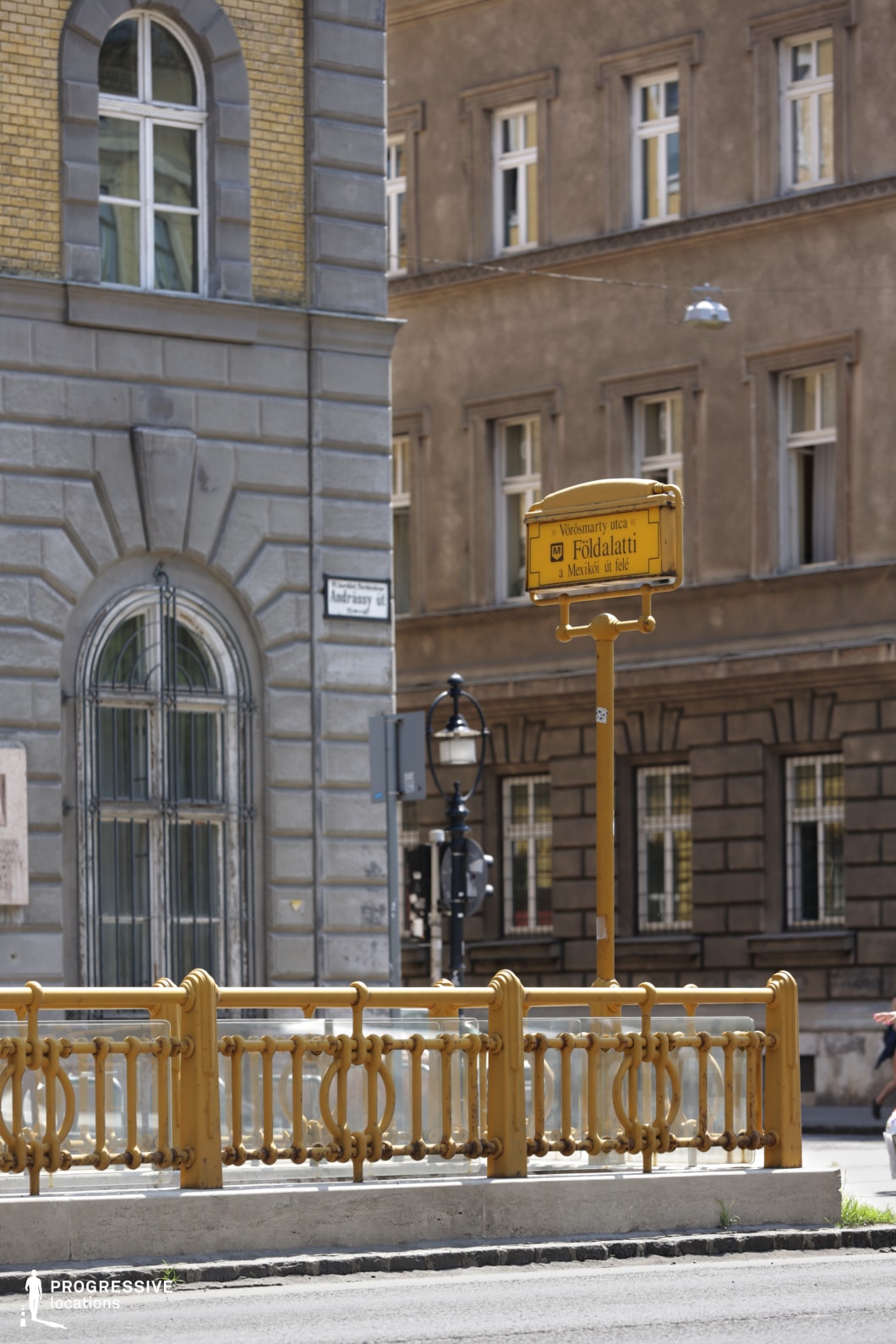 This image captures a historic metro entrance framed by elegant yellow cast-iron railings and a matching vintage-style signpost. The detailed metalwork and signage reflect a classic design, suggesting an early underground transit system with a strong architectural identity. In the background, ornate historic buildings with arched windows, decorative stonework, and muted tones provide a contrast to the bright yellow metro elements. A traditional street lamp further enhances the old-world charm of the scene. The combination of classic urban infrastructure and refined architecture makes this an ideal location for historical or atmospheric cityscape compositions.