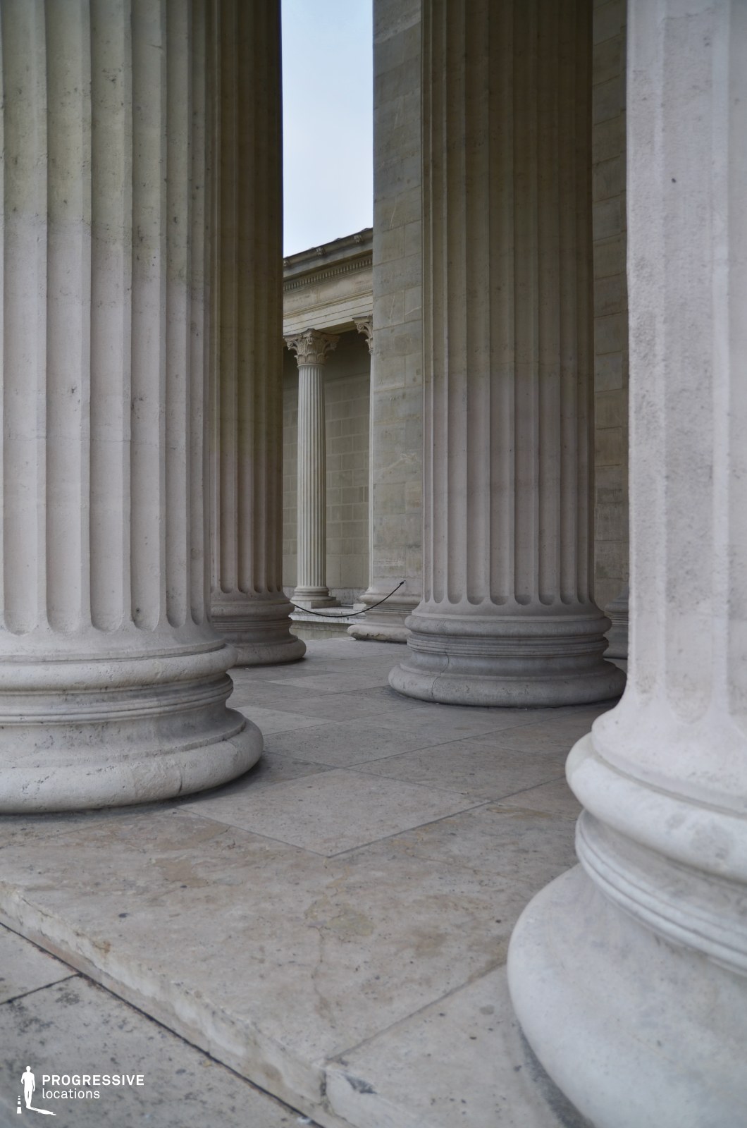 This image showcases a grand neoclassical colonnade with massive, fluted limestone columns arranged in a rhythmic sequence. The classical architectural elements, including the sturdy bases and carved capitals, emphasize a sense of permanence and grandeur. The perspective between the columns creates a layered depth, leading the eye toward an elegant entablature in the background. The smooth stone flooring complements the stately composition, reinforcing the historic and institutional character of the space. This setting is ideal for cinematic compositions requiring a formal, monumental backdrop, particularly for themes of power, history, or academia.
