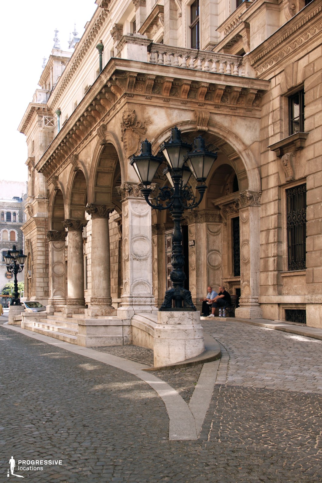 This image captures the elaborate entrance of a historic opera house, featuring grand arches supported by intricately carved stone columns. The building showcases classical and baroque architectural details, including sculpted reliefs, decorative cornices, and finely crafted balustrades. A prominent black wrought-iron street lamp with an ornate design stands in the foreground, enhancing the old-world charm of the scene. The cobblestone street and gently curving pathways add to the European atmosphere, making this an ideal setting for cinematic, historical, or sophisticated urban visuals. The composition emphasizes elegance, grandeur, and architectural craftsmanship.