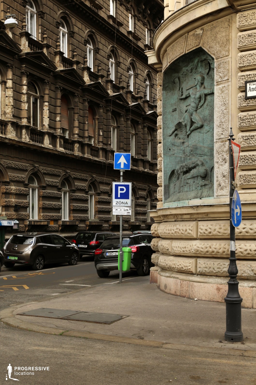 This image captures a European street corner framed by two grand historic buildings with richly ornamented facades. The architecture features intricate stone detailing, arched windows, and textured surfaces, exuding a classical and slightly aged character. A prominent bronze relief is embedded into the curved corner of one building, depicting a dynamic sculptural scene that adds an artistic focal point. The street is lined with parked cars, and urban elements such as signage and wrought-iron lampposts contribute to the authentic city atmosphere. The strong contrast between the dark masonry and lighter stone highlights the depth and texture of the space, making it an ideal setting for urban documentary, historical narratives, or atmospheric cityscape photography.