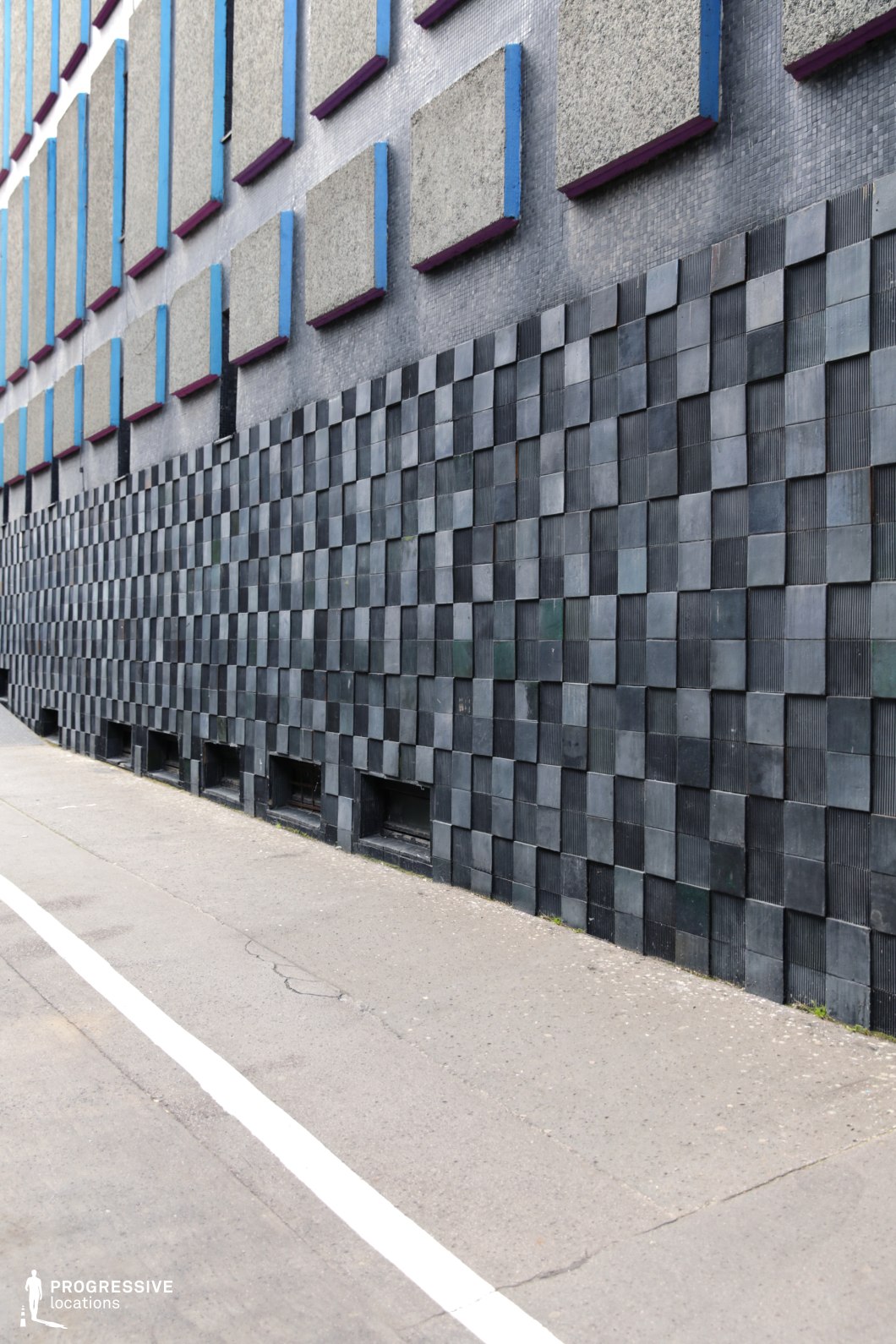 This image features a striking brutalist or modernist architectural facade, distinguished by its geometric and textured surface treatments. The lower portion of the wall is clad in dark, offset square tiles, creating a three-dimensional effect, while the upper section consists of protruding concrete panels with blue and purple edging. The contrasting materials and structured repetition give the space a raw, industrial aesthetic. The sidewalk and road markings further emphasize the stark, urban character of the setting. This location is ideal for contemporary, dystopian, or architectural-themed visual projects, highlighting minimalist and socialist-era design influences.